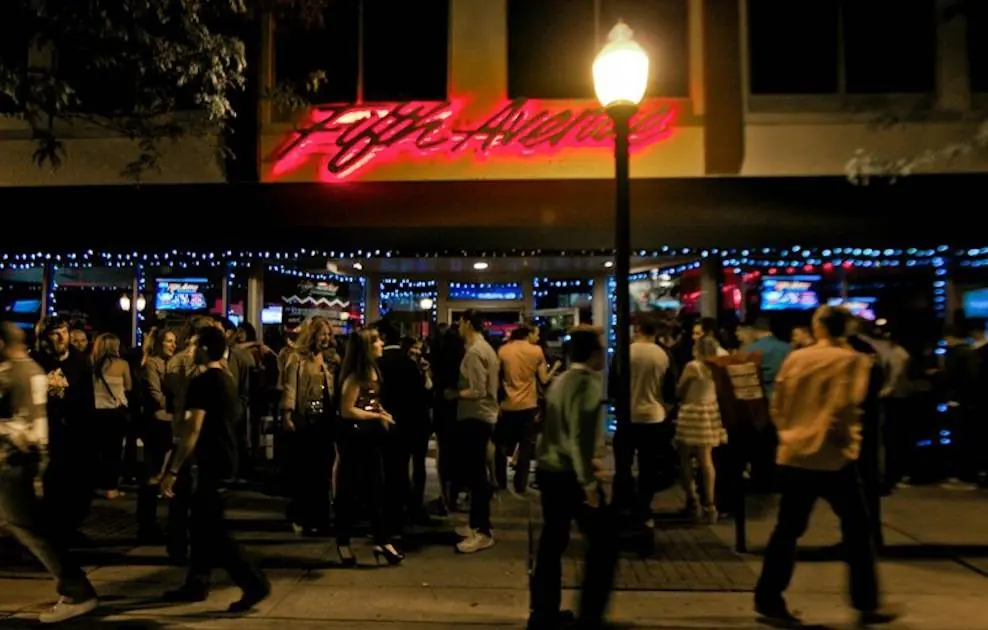 Fifth Avenue (Royal Oak) at night: red neon 'Fifth Avenue' script sign, string lights and a crowd of people gathered outside under a street lamp.