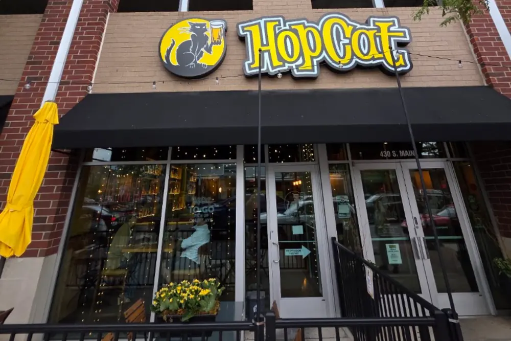HopCat storefront in daylight: yellow-and-black HopCat cat logo sign over a black awning and glass doors, red-brick facade, yellow patio umbrella.